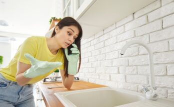 A woman wearing green latex gloves, talking on the phone while leaning on the kitchen counter, looking at the sink.