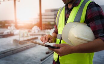 An engineer on a construction site with a safety vest on and a hard hat in his hands. He is looking at a tablet.