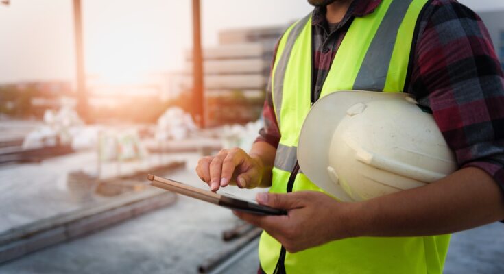 An engineer on a construction site with a safety vest on and a hard hat in his hands. He is looking at a tablet.
