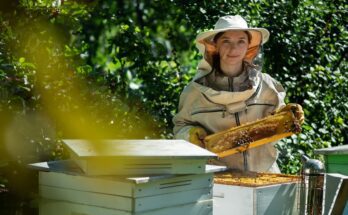 A woman in a beekeeping suit standing outdoors near beehives, holding up a frame filled with honeycomb, honey, and bees.