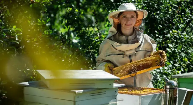 A woman in a beekeeping suit standing outdoors near beehives, holding up a frame filled with honeycomb, honey, and bees.