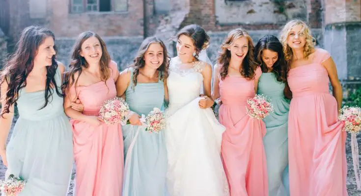 A bride in a wedding gown standing beside six women wearing long pink or blue dresses, each holding a bouquet of flowers.