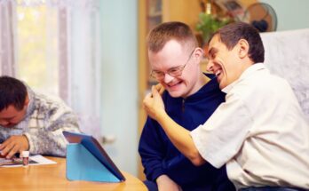 Two adults in a developmental home hugging and smiling while another person writes at a nearby table.