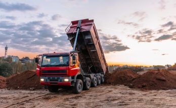 A red dump truck unloading dirt onto a heap on the ground as the sun sets under a partly cloudy sky.