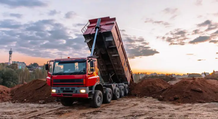 A red dump truck unloading dirt onto a heap on the ground as the sun sets under a partly cloudy sky.