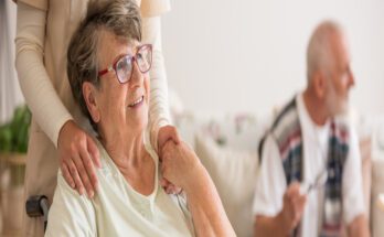 An aging woman sits in a wheelchair at her new care facility as her adult child pushes the chair. She's smiling.