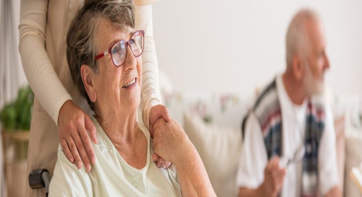 An aging woman sits in a wheelchair at her new care facility as her adult child pushes the chair. She's smiling.