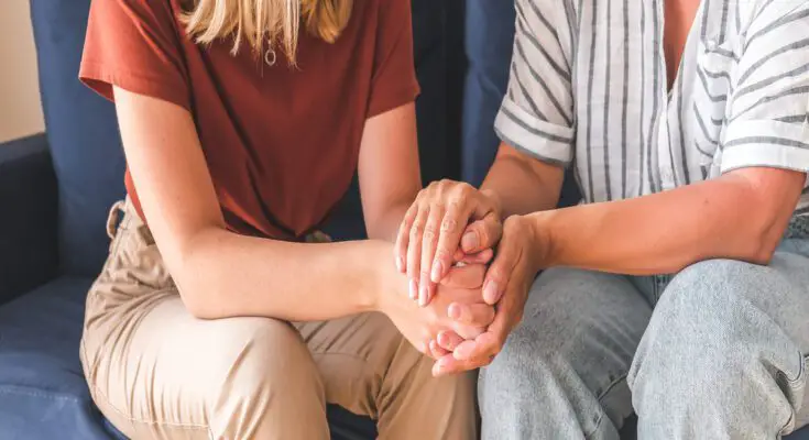 A close-up of two people from the neck down sitting next to one another on the couch. They hold their hands together.