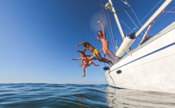 A group of young adults leap off the side of a white boat into calm ocean waters under a clear blue sky.