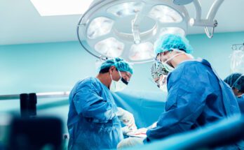 A surgical team in blue scrubs and masks operates under a bright overhead light in a sterile operating room.