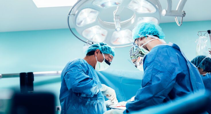 A surgical team in blue scrubs and masks operates under a bright overhead light in a sterile operating room.