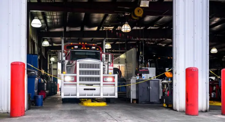 A red and white semi-truck sits parked in a well-lit workshop equipped with repair tools and equipment.
