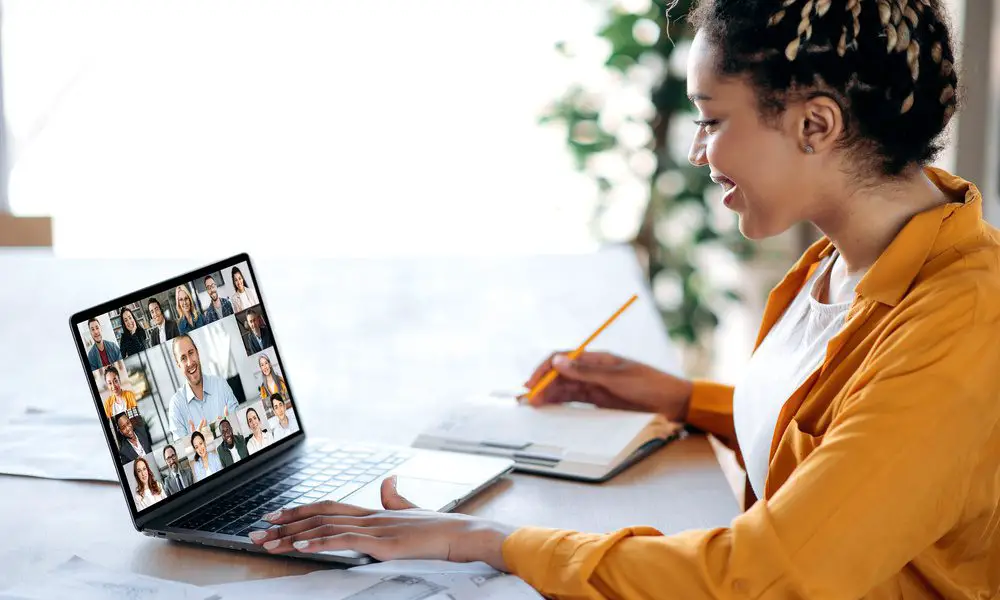 A woman sits in her office, looking at the screen on her laptop, where she is telecommuting with multiple people.