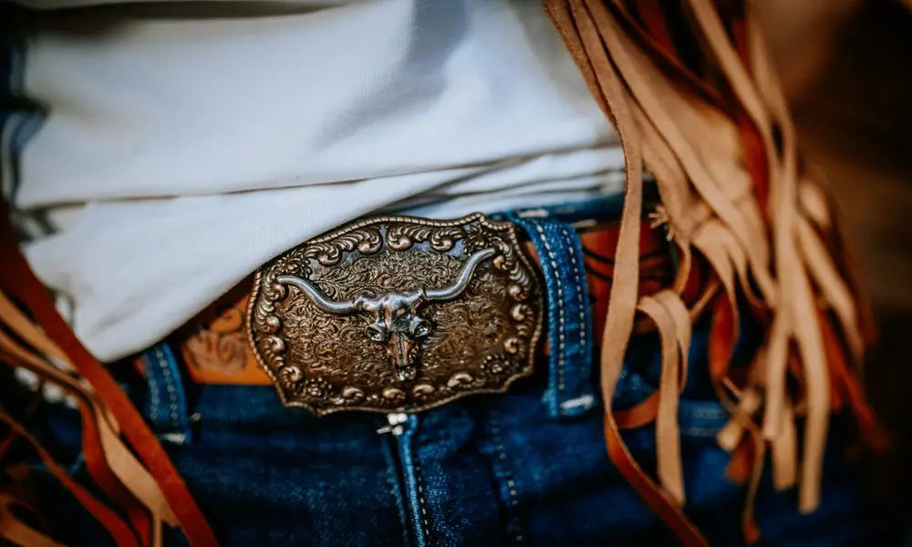 A person wearing a white shirt, a tassel vest, and jeans cinched by a leather belt with an ornate western-style buckle.