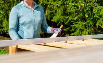 A woman wearing a teal zip-up jacket using a brush to coat a wooden pallet with white paint outdoors.