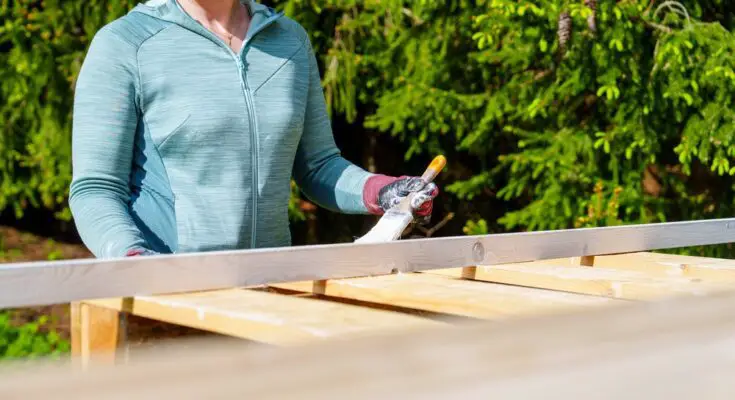 A woman wearing a teal zip-up jacket using a brush to coat a wooden pallet with white paint outdoors.
