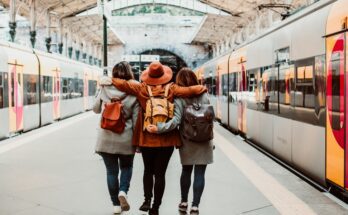 Three people walk through a train station with their arms wrapped around each other. Trains wait on either side of the platform.