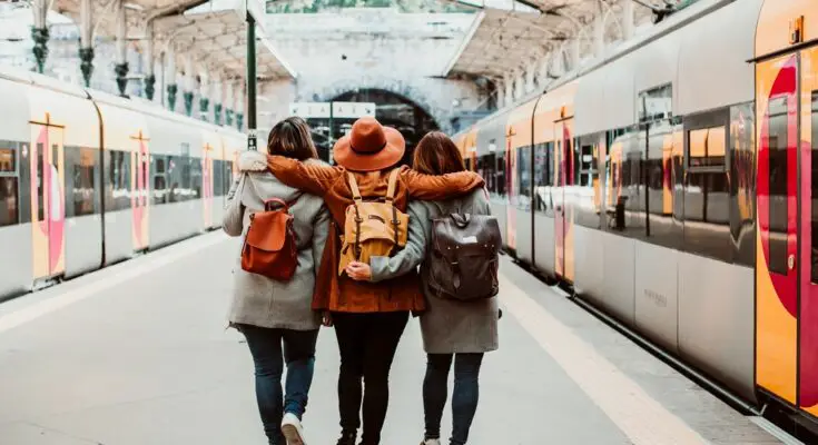 Three people walk through a train station with their arms wrapped around each other. Trains wait on either side of the platform.