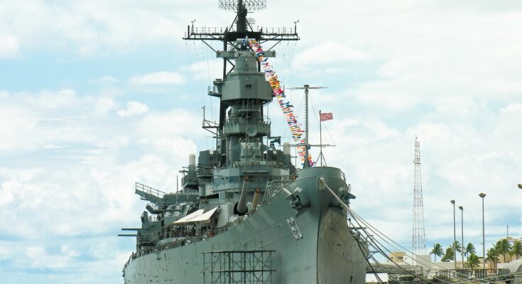 A towering gray battleship with a with the number "63" printed on its side sitting in quiet, calm water at a harbor.
