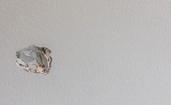A close-up of a white, slightly textured wall with a small hole partially smashed into the left side near the middle.