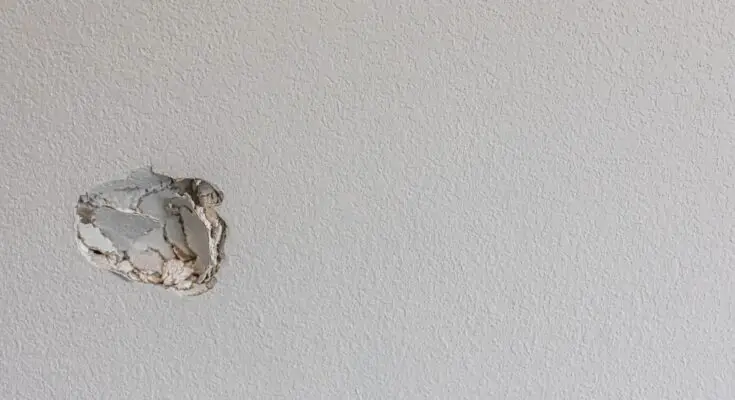 A close-up of a white, slightly textured wall with a small hole partially smashed into the left side near the middle.