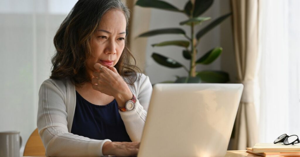 An older woman sits at a wooden table looking at a laptop. She is rubbing her chin with one of her hands.