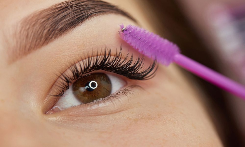 An extreme close up of a woman's brown eye with a purple spoolie held up to the curled, black eyelashes.