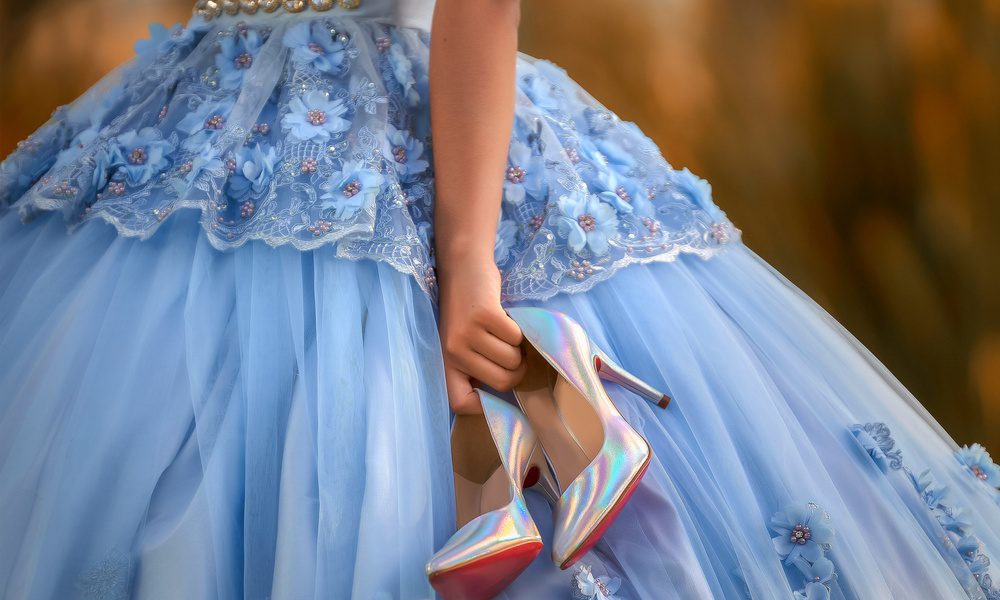 A partly seen young woman in a blue quinceañera gown holding iridescent high heels beside her skirt.