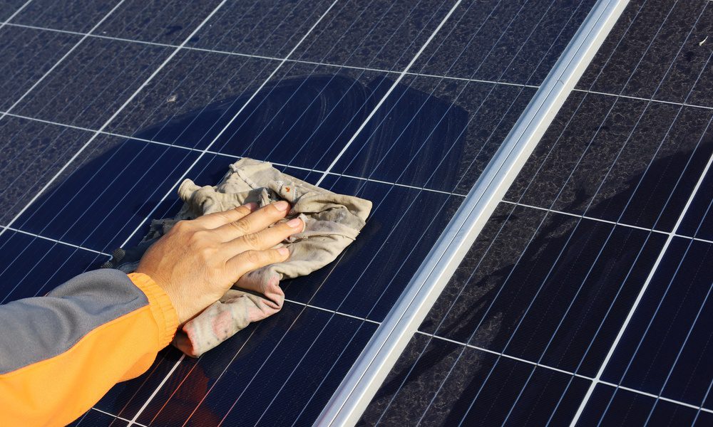 A person's hand is cleaning a solar panel with a towel. Some of the panels are clean, while other parts still have dust.