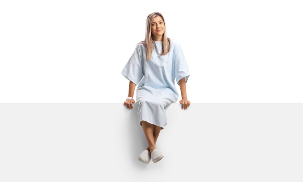 A woman in a hospital gown sitting on a white blank panel with legs crossed against a white background.