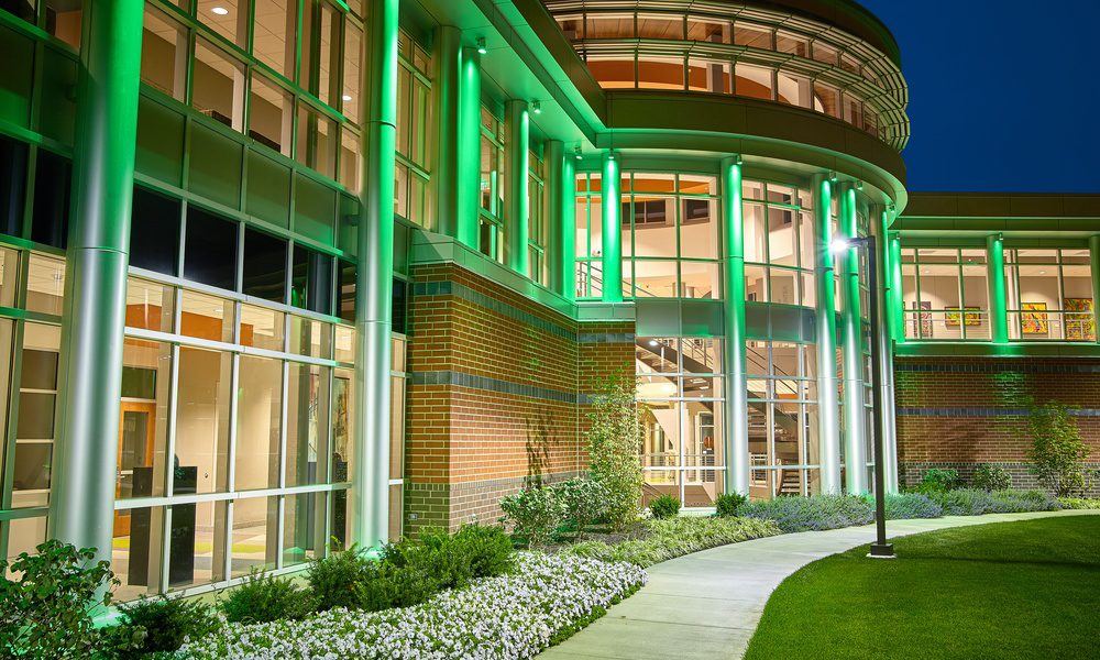 A green-lit brick and glass building at night with tall columns, large windows, a curved walkway, lawn, and flower beds.