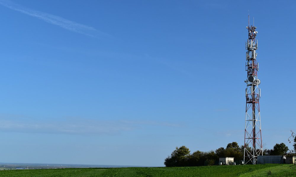 A telecommunication tower with antennas rests in the top part of a lush, green field. Small buildings surround the tower.