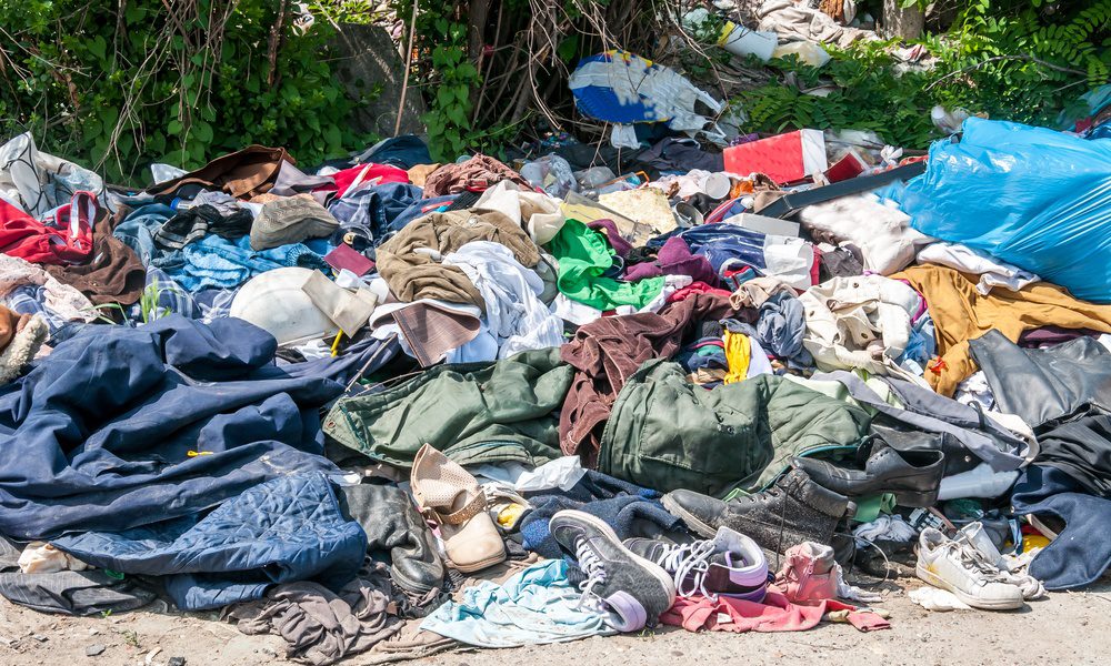 A large pile of shoes, shirts, and pants on a sandy surface. Trees and greenery are in the background.