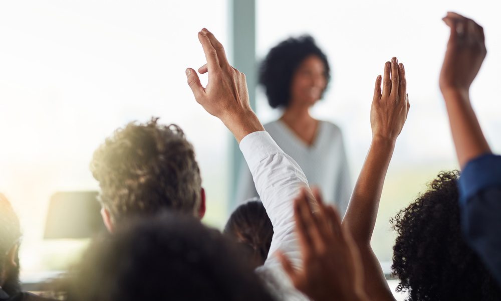 Several people raise their hands to ask questions during a business meeting. A manager stands in front of them.