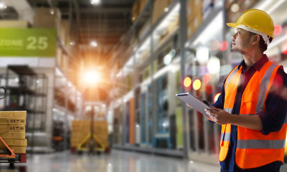 A manager walks through a warehouse, wearing a hard hat and an orange vest. He refers to a tablet as he walks.