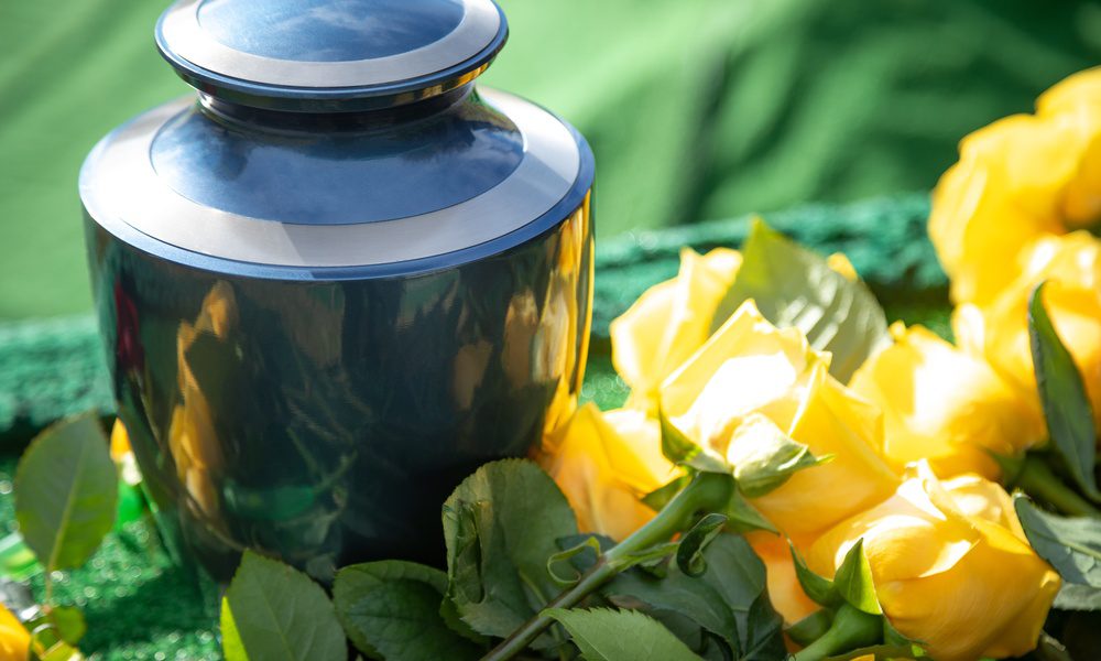 A dark metal urn resting on green fabric beside yellow roses and green leaves in bright outdoor light, with soft greenery.