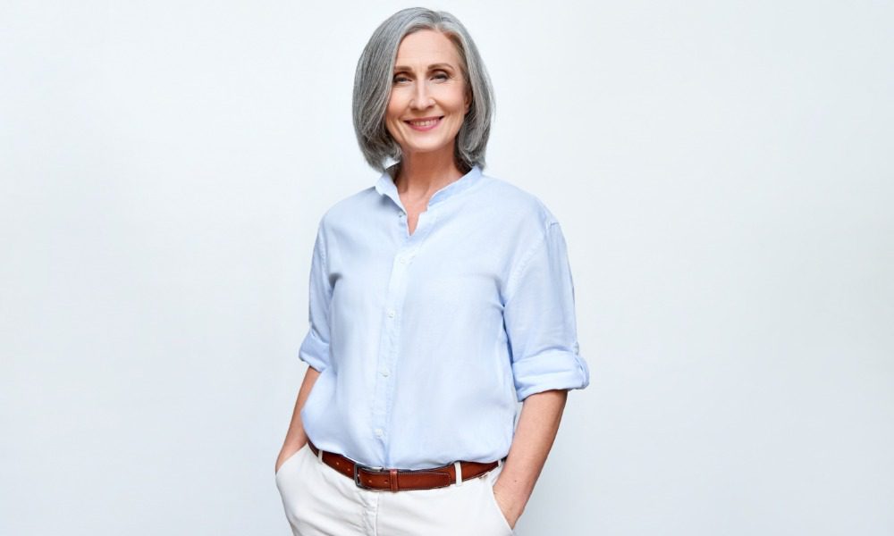 A woman with shoulder-length gray hair stands with her hands in her pockets. She wears a blue shirt and white pants.