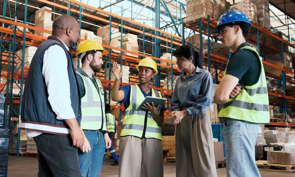 Five workers wearing safety vests and hard hats meeting with a tablet between tall warehouse racks and boxes.