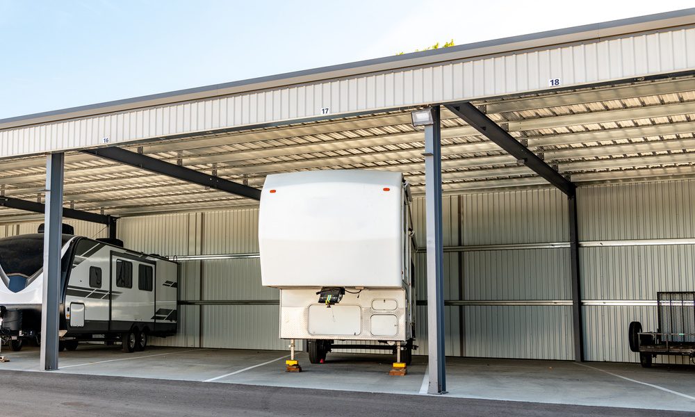 A white RV trailer parked beneath a metal-covered storage bay with another trailer in the next bay over.