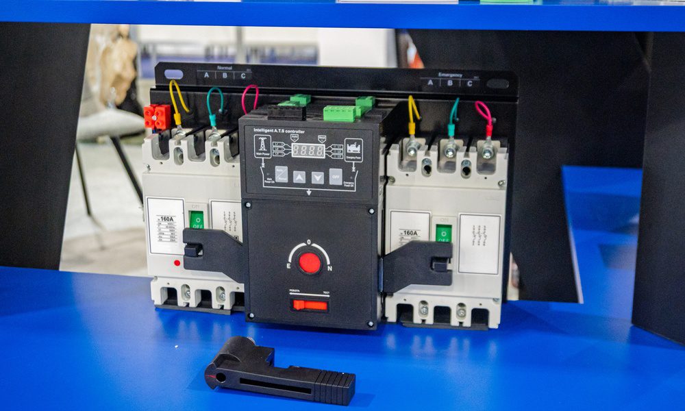 A long rectangular automatic transfer switch with a red button sits on a blue table in a large brightly lit space.