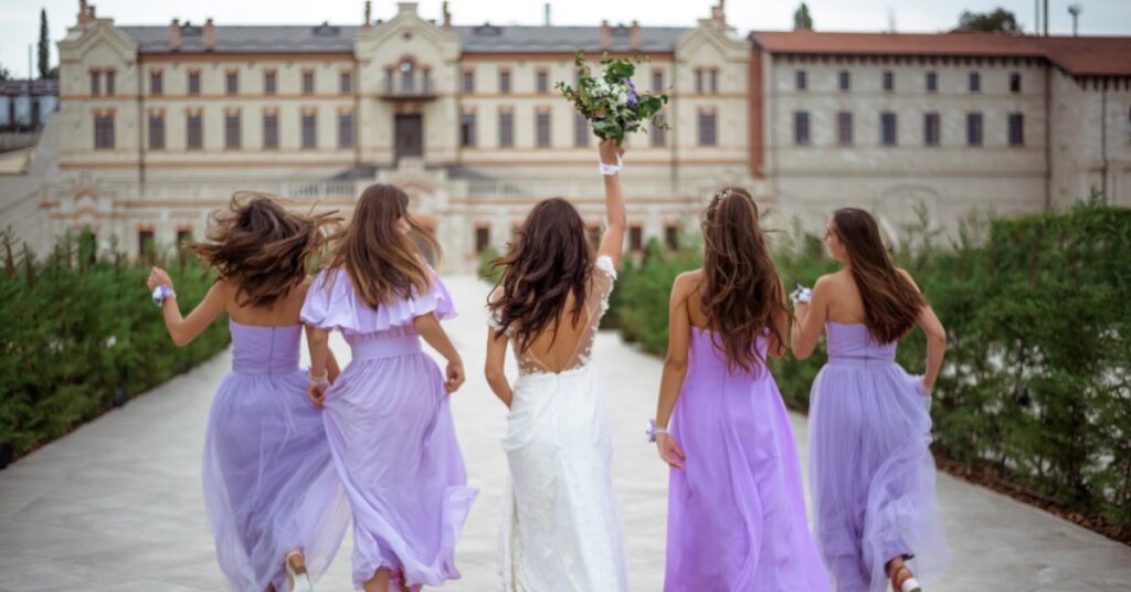 A bride and four women in lavender dresses running along a walkway, with the bride holding up a bouquet.