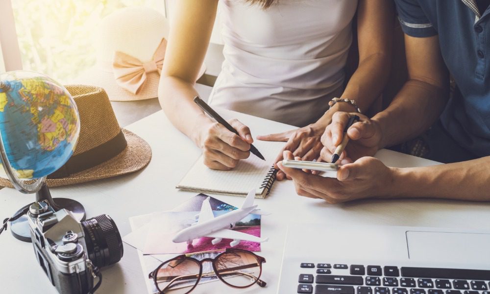 A couple sits at the table together to plan an international trip. There's a laptop, a camera, a globe, and they're writing.