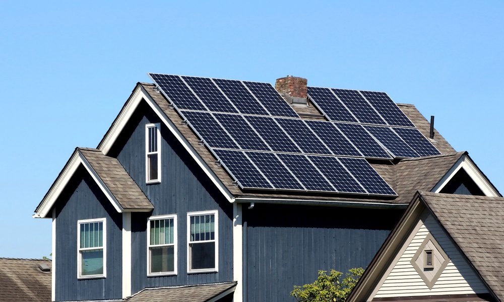 Solar panels cover the roof of a dark blue two-story house with white trim beneath a clear blue sky.
