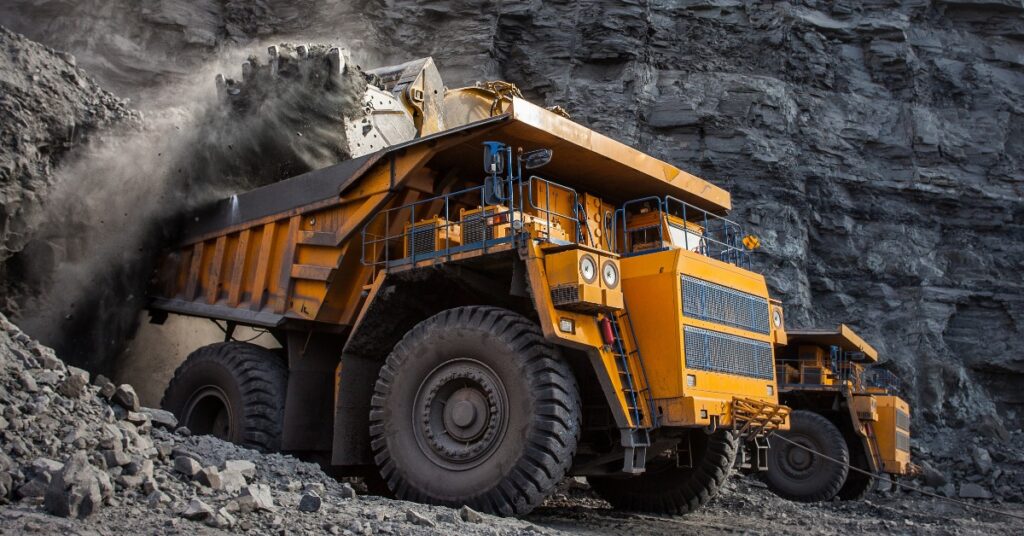 A large yellow mining truck being loaded with dark coal beside a rocky wall in an open coal mine, with dust in the air.