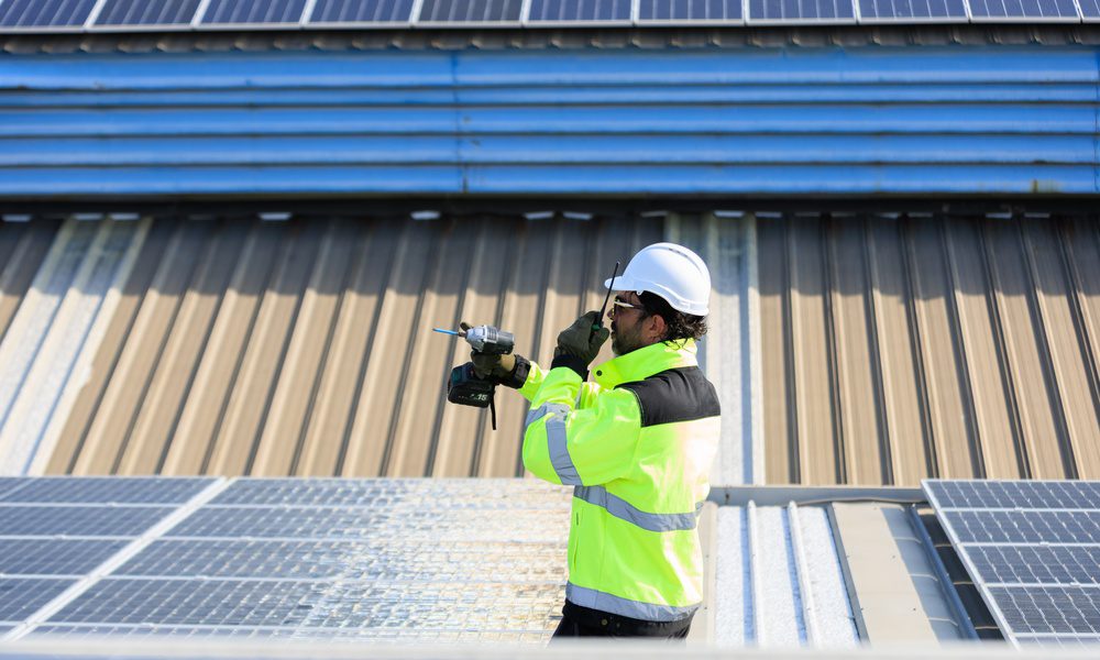 Solar technician in a safety helmet uses a power drill and handheld radio while working among rooftop solar panels.