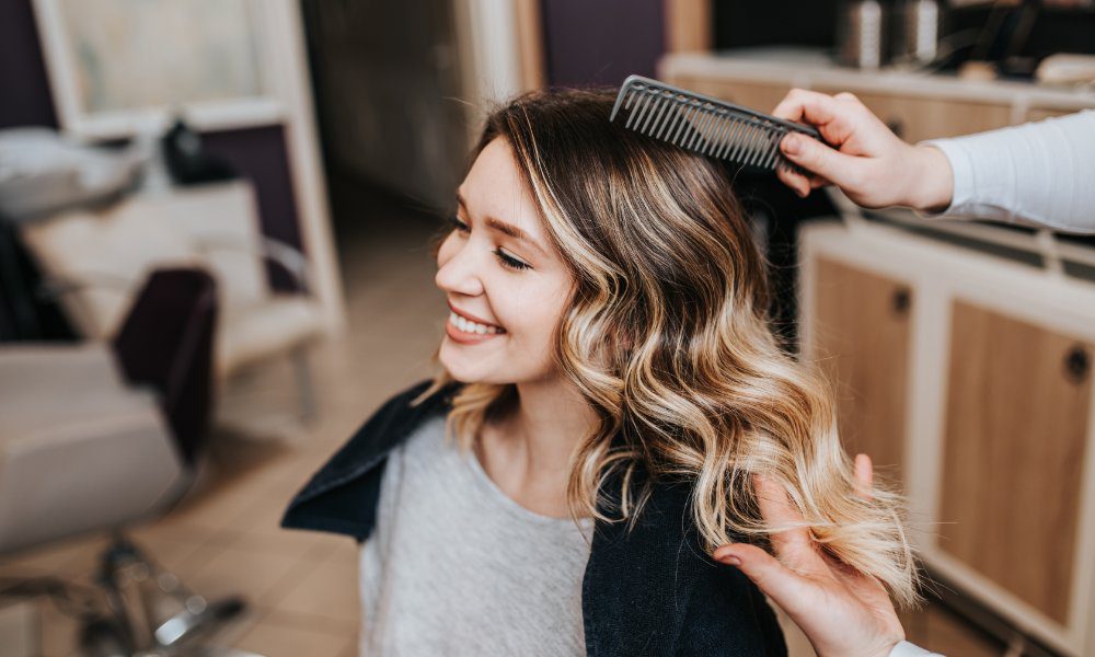 Stylist uses a wide-tooth comb while arranging the highlighted waves of a smiling woman seated in a hair salon.