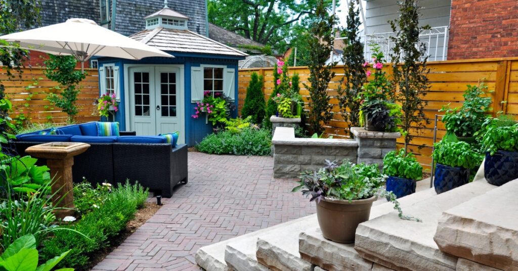 A backyard surrounded by a wooden fence, with multiple potted plants, an outdoor sectional couch, and a large umbrella.