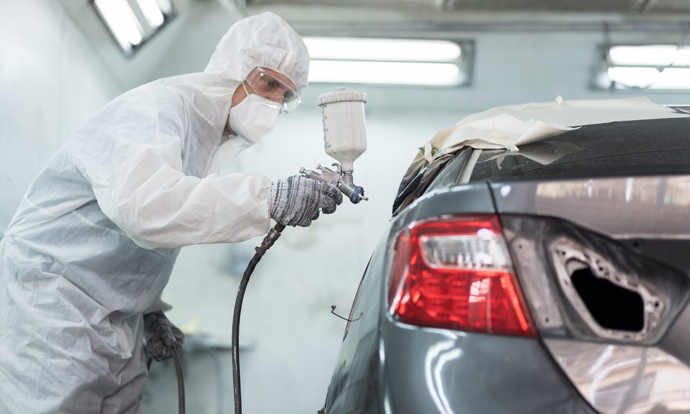 A man wearing a white protective suit, protective glasses, and a mask is using a sprayer to paint a car gray.