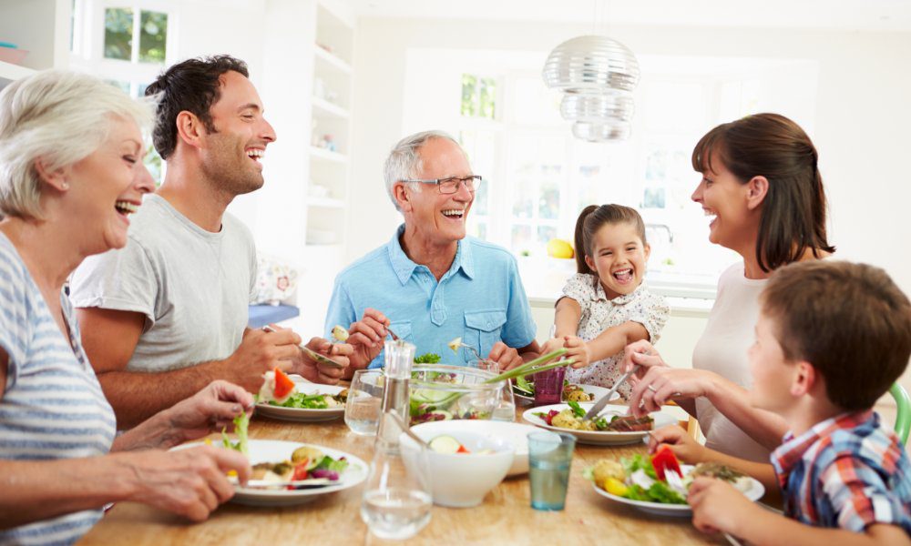 A multigenerational family sits around a dining table, smiling and laughing together while sharing a meal.