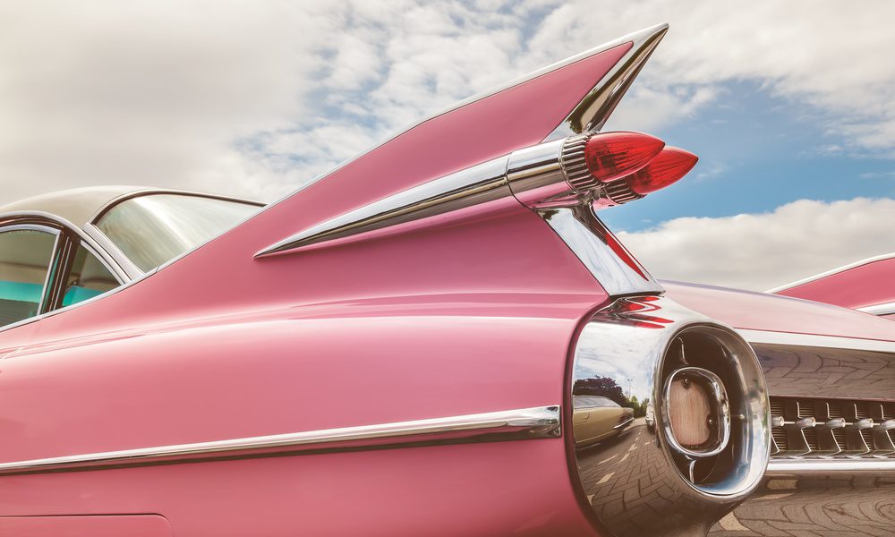 Rear detail of a pink vintage car showcasing chrome tail fins, circular taillight housing, and bright blue sky with clouds.